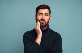 Dental problems. Portrait of unhealthy man pressing sore cheek, suffering acute toothache, periodontal disease, cavities or jaw pain. Indoor studio shot isolated on blue background